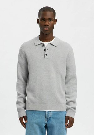 Man wearing light gray knit polo sweater with black buttons and light blue jeans, standing against a plain white background.