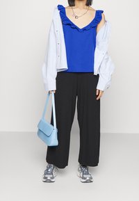 Débardeur bleu à volants, chemise ample bleu clair, pantalon noir à jambes larges, sac à main bleu texturé, et baskets grises avec détails métalliques.