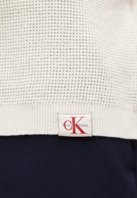 Ourlet de pull en maille texturé crème avec une étiquette en tissu blanc affichant le logo rouge "cK" et le texte gris "Calvin Klein" au-dessus d'un pantalon bleu marine.