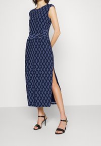 Robe sans manches bleu marine avec un motif en losanges, taille cintrée et fente latérale. Associée à des sandales à talons hauts noires à lanières.