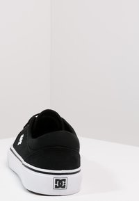 Black canvas skate shoe with a low profile, white rubber sole, and a prominent white logo on the heel. Flat laces and stitched accents.