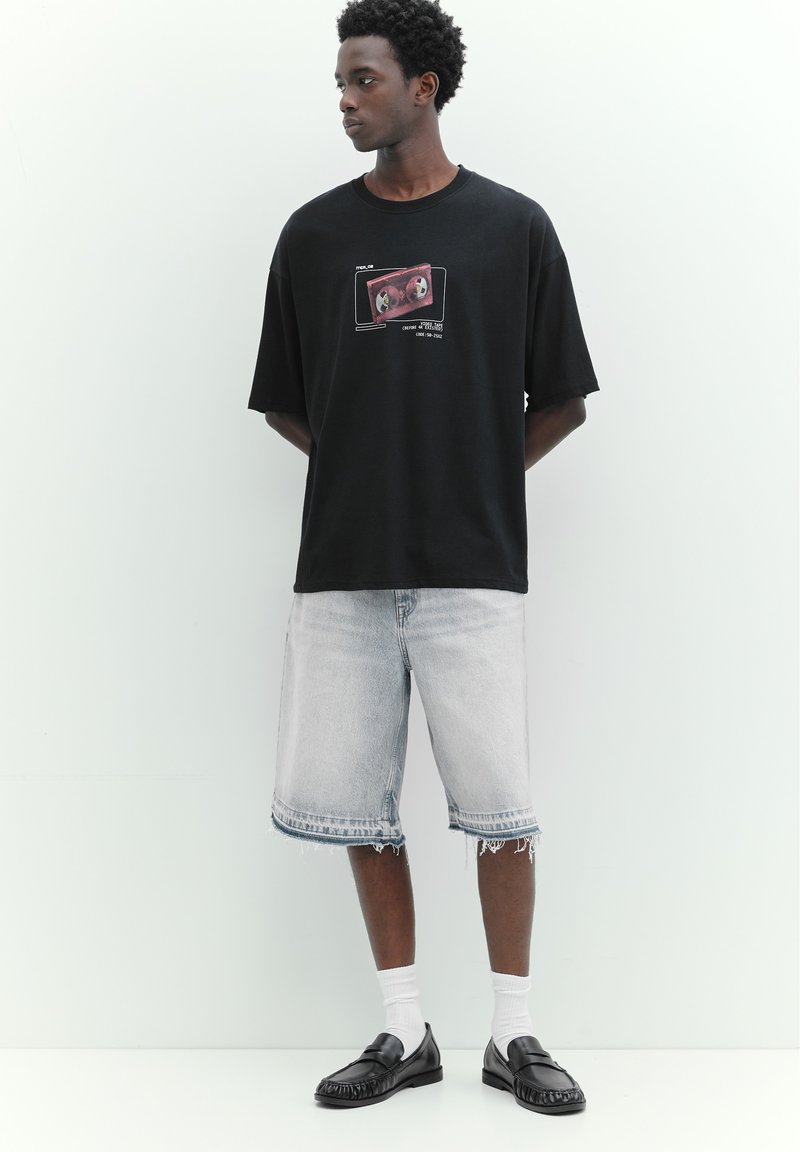 Jeune homme portant un t-shirt noir avec un motif graphique, un short en jean clair, des chaussettes blanches et des mocassins noirs, debout devant un fond clair uni.