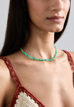 Collier en perles turquoise avec des pierres de forme irrégulière, porté sur un top en crochet brun-rouge, mettant en valeur une texture de peau lisse.