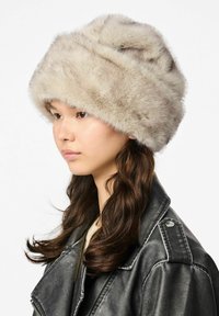 Lažni krzneni šešir u svijetlo bež boji s mekanom, pahuljastom teksturom, koja ima široki, preklopljeni dizajn i istaknute detalje oko oboda.