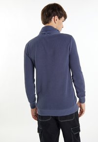 Marineblaues Strickpullover mit strukturiertem Muster, Schalkragen und gerippten Bündchen. Über schwarzen Cargo-Hosen getragen, von hinten gezeigt.