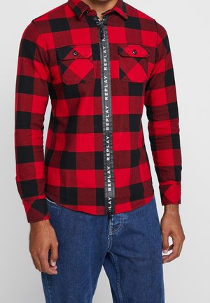 Homme portant une chemise à manches longues à carreaux rouge et noir avec deux poches poitrine à boutons et une fermeture éclair noire étiquetée "REPLAY", associée à un jean bleu.