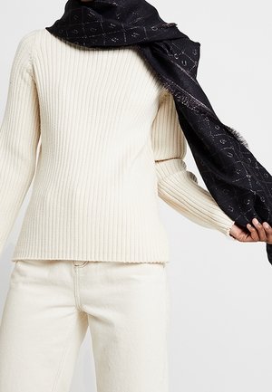 Personne portant un pull crème côtelé, un pantalon assorti, et tenant une écharpe noire à motifs drapée sur une épaule.