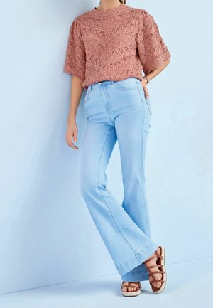 Kobieta ubrana w krótki sweter w kolorze pudrowego różu z wzorem, jasnoniebieskie, dzwoniaste, z wysokim stanem dżinsy oraz brązowe sandały na paski.