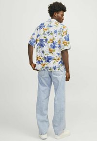 Kurzarmhemd mit weißem Hintergrund, das ein farbenfrohes Blumenmuster in Blau, Gelb und Grün zeigt. Getragen mit hellblauen Jeans.