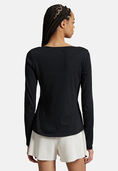 Femme aux cheveux tressés portant un haut noir à manches longues et un short blanc, debout de dos devant un fond uni.