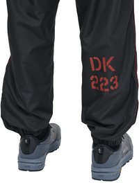 Underben iført sorte bukser med rød tekst "DK 223" og grå sneakers med sorte hælflipper, set bagfra mod en hvid baggrund.