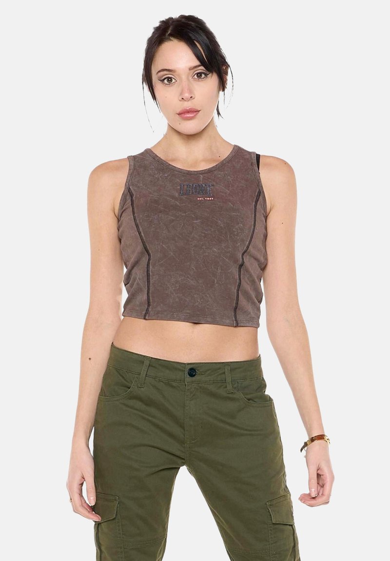 Jeune femme aux cheveux foncés porte un crop top marron sans manches et un pantalon cargo vert olive, debout devant un fond uni.