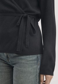 Schwarze langärmlige Wickeljacke, die an der Taille gebunden ist, kombiniert mit verblassten blauen Jeans auf einer Person vor einem schlichten Hintergrund.