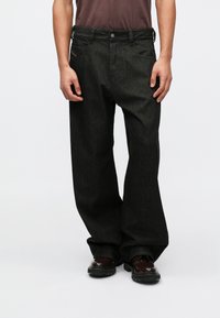 Calças de ganga pretas com um corte descontraído, apresentando um design de cinco bolsos, passantes para cinto e detalhes de costura discretos; usadas com sapatos pretos.