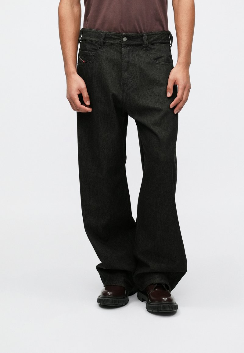 Calças de ganga pretas com um corte descontraído, apresentando um design de cinco bolsos, passantes para cinto e detalhes de costura discretos; usadas com sapatos pretos.