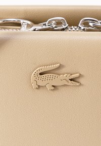 Μπεζ δερμάτινο αντικείμενο με ανάγλυφο λογότυπο κροκόδειλου και εμφανείς ασημένιους μηχανισμούς φερμουάρ στην κορυφή.