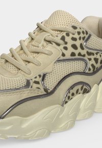 Baskets beiges en mesh et suède, avec des accents imprimé léopard, semelle épaisse rainurée, et lacets beiges sur fond blanc.