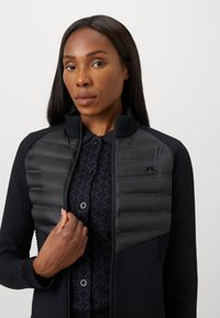 Mulher a usar uma jaqueta acolchoada preta com fecho éclair sobre uma camisa de botão estampada, segurando o fecho da jaqueta com uma mão contra um fundo simples.