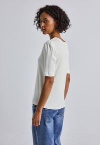Haut blanc à manches courtes avec des manches bouffantes et un col rond, associé à un jean en denim bleu clair, présenté sous un léger angle latéral.