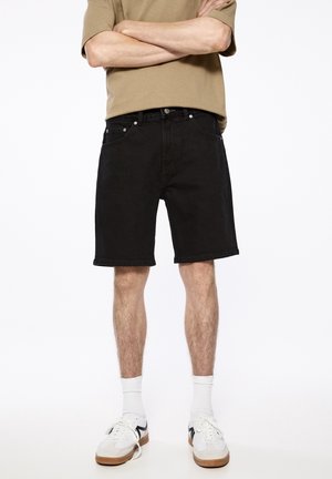 Black denim shorts with a straight cut, five pockets, and a subtle semi-matte finish. Worn with a tan shirt and white athletic shoes.