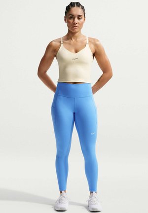 Femme debout les mains dans le dos, portant un haut de sport beige, des leggings bleu vif et des baskets blanches sur un fond uni.
