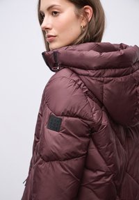 Bordeauxfarbiger Puffer-Jacke mit hohem Kragen, gestepptem Design und einem schwarzen Logo-Patch auf der Schulter. Verfügt über Reißverschluss-Beschläge und Kapuze.