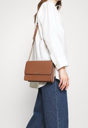 Kobieta ubrana w białą bluzkę i niebieskie dżinsy, niosąca brązową skórzaną torbę przez ramię z złotym łańcuszkiem na pasku.