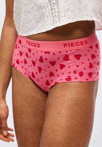 Γυναίκα που φοράει ροζ εσώρουχα σε στυλ hipster με κόκκινα σχέδια καρδίας και την επιγραφή «PIECES» στη μέση, συνδυασμένα με ένα λευκό crop top με κέντημα eyelet.