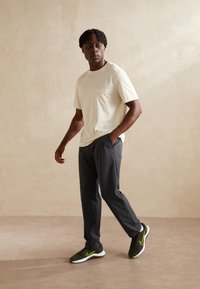 Helles Cremefarbenes T-Shirt mit kurzen Ärmeln und Brusttasche, kombiniert mit dunkelgrauen, schmal zulaufenden Hosen und schwarzen Sneakern mit gelben Akzenten.