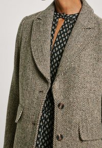 Beżowo-brązowy żakiet w wzór houndstooth wykonany z mieszanki wełny, posiadający klapki, dwie przednie kieszenie oraz zapięcie na guziki.