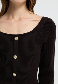 Zwart geribd gebreid top met een vierkante halslijn en drie decoratieve gouden knopen aan de voorkant, met een zachte textuur en subtiele glans.