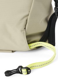 Eckdetails des beigen Stoff-Rucksacks zeichnen sich durch eine strukturierte Oberfläche, verstärkte Nähte und einen leuchtend gelben Riemen mit reflektierenden Akzenten sowie einem schwarzen Clip aus.