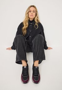 Giacca oversize in denim nero, jeans neri a gamba larga e sneakers nere e borgogna con dettagli texture, seduto su uno sfondo bianco.