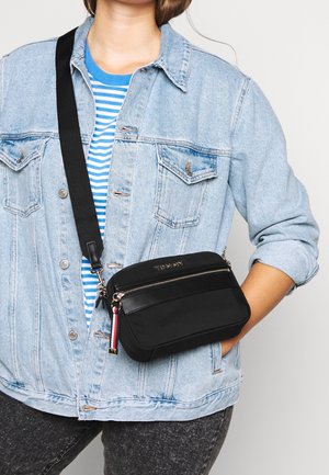 Persoon draagt een licht denim jack en een blauwgestreept shirt met een zwarte crossbodytas met een ritssluiting aan de voorkant en een merkdetail.