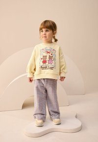 Żółta bluza z nadrukiem misia, z bufiastymi rękawami, zestawiona z brązowymi spodniami w kratkę. Dziecko ma na sobie beżowe trampki i stoi na zakrzywionej platformie.