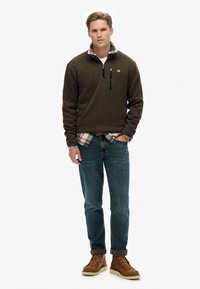 Bruine fleecetrekker met halve rits, gedragen over een geruit shirt. Gemaakt met blauwe spijkerbroeken en bruine veterschoenen.
