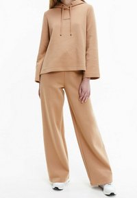 Sweatshirt à capuche marron avec logo, coupe décontractée et manches longues, associé à un pantalon large assorti en tissu doux. Des baskets blanches complètent le look.