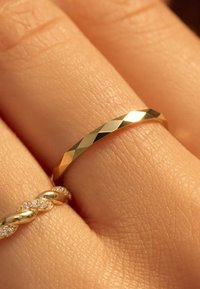 Bague en or avec un design facetté, présentant des bords plats et une surface lisse, présentée sur une main aux côtés d'un anneau en or torsadé.