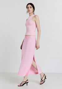 Helles rosa Zweiteiler-Outfit, bestehend aus einem taillierten ärmellosen Oberteil und einem langen Rock mit seitlichen Schlitzen und einem Kordelzugbund. Schwarze Riemchen-Absätze.