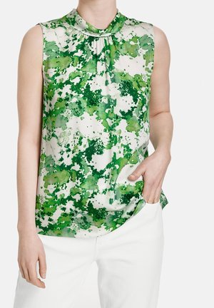 Haut sans manches avec un motif abstrait aquarelle vert et blanc, porté avec un pantalon blanc, la main reposant dans la poche.