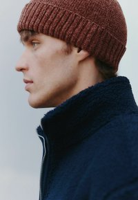 Brun ribbet hue, marineblå sherpa zip-up jakke med høj krave. Fokus på teksturerede materialer og kontrasterende farver.