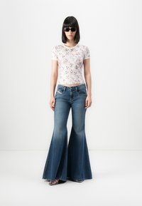 Kvinde iført sorte solbriller, en hvid tætsiddende t-shirt med katteansigt og højtaljede blå flared jeans, stående foran en hvid baggrund.