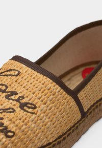 Chaussure beige tissée sans lacets avec des bordures marron et un texte brodé en brun foncé sur la partie supérieure, dotée d'une semelle plateforme texturée.
