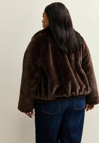 Una donna con lunghi capelli scuri indossa una giacca di pelliccia sintetica marrone scuro e jeans blu scuro, in piedi di spalle contro uno sfondo chiaro e semplice.