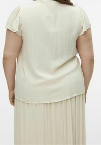 Blouse crème clair avec des manches courtes froncées, présentant une texture lisse et un ourlet arrière légèrement plus long. Associée à une jupe plissée.