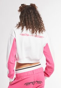 Felpa corta bianca con maniche rosa, con grafiche di fiamme sul retro. Abbinata a pantaloni rosa, accentuati da un design nero.