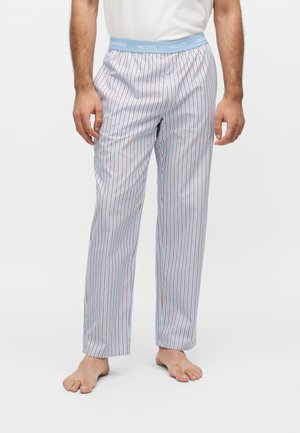Homme portant un pantalon rayé vertical bleu clair et blanc avec une ceinture bleue marquée Lacoste, debout pieds nus sur un fond uni.