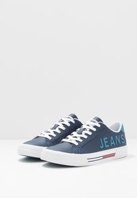 Blue sneaker with white laces, featuring "JEANS" in teal on the side, smooth leather upper, and textured rubber sole with a striped accent.