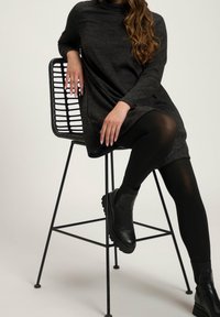 Dark gray, long-sleeve dress made of soft fabric, layered over black tights and paired with black ankle boots. Model seated on a wire chair.
