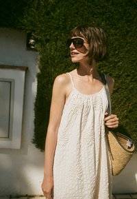 Femme en robe blanche sans manches et grandes lunettes de soleil tenant un sac en paille devant des arbustes verts et un mur blanc.